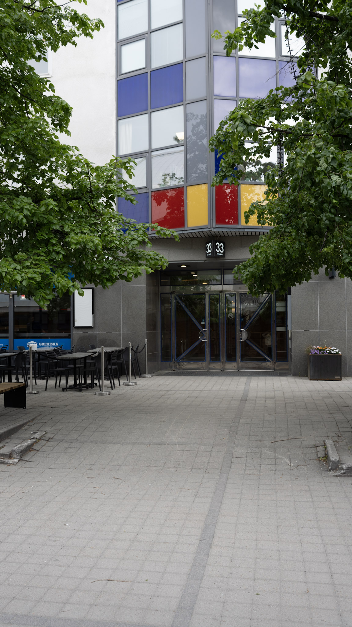 Entré till en byggnad med glasdörrar och färgade fönster, omgiven av gröna träd.